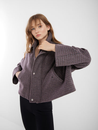 Eine Frau posiert selbstbewusst vor einem schlichten Hintergrund und trägt die Jacke Victoria mit Hahnentrittmuster, weiten Ärmeln und schwarzer Hose. Sie lüftet den Kragen dieser stilvollen Übergangsjacke und blickt mutig in die Kamera.