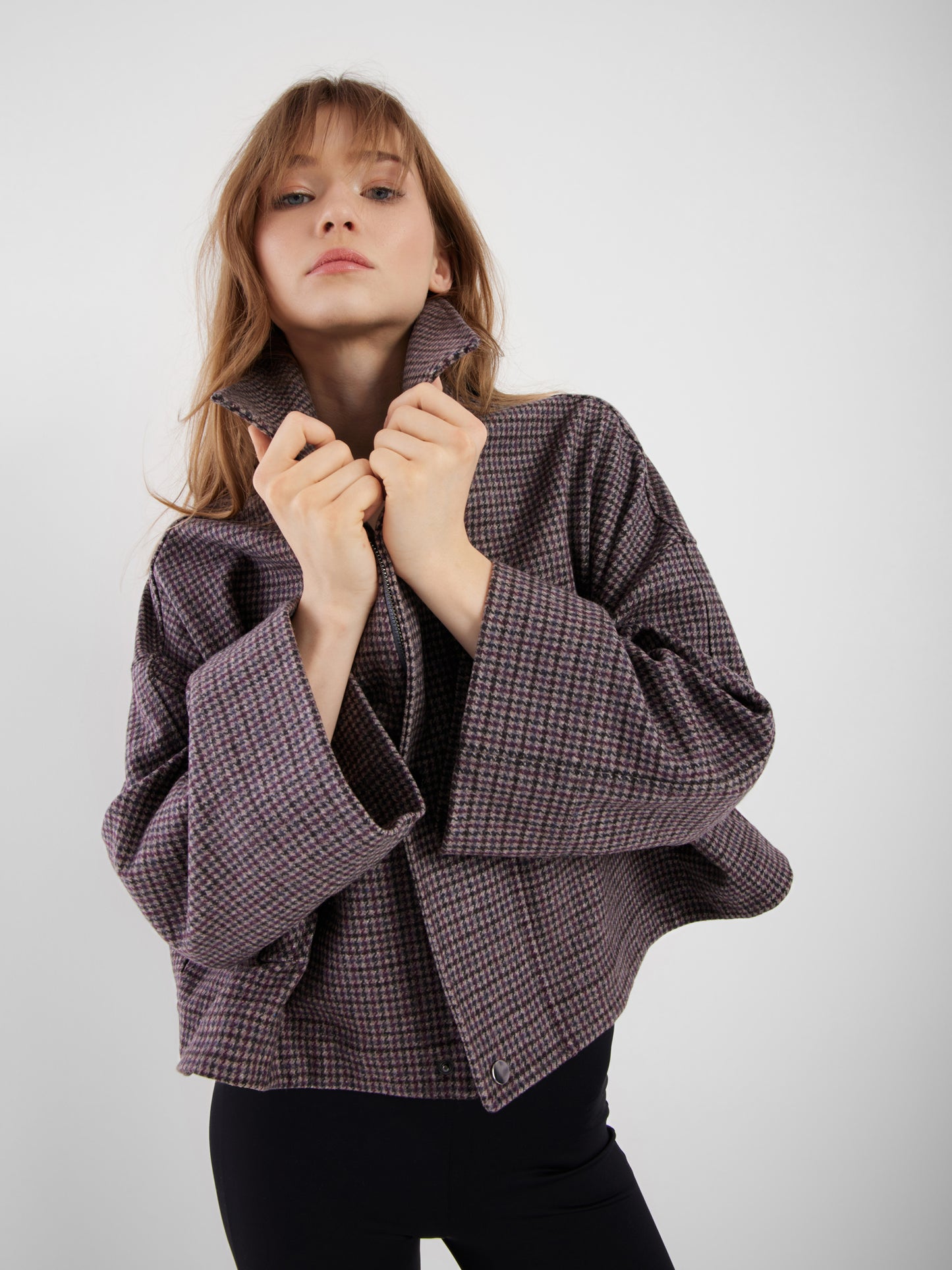 Eine junge Frau mit langem, hellbraunem Haar posiert vor einem schlichten Hintergrund und trägt die lockere Jacke Victoria mit Hahnentrittmuster und weiten Ärmeln sowie eine dunkle Hose. Sie hält den Kragen fest und blickt selbstbewusst in die Kamera.
