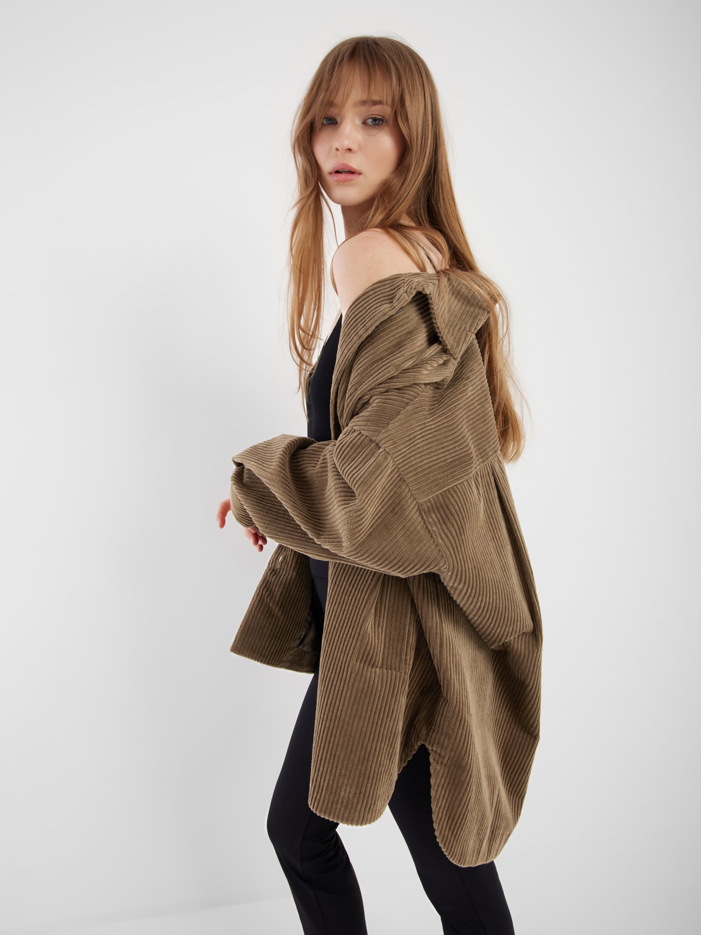 Eine Frau mit langem, hellbraunem Haar trägt den Mantel Ted Khaki Coderoy über einer Schulter über einem schwarzen Outfit, steht vor einem schlichten weißen Hintergrund und blickt in die Kamera.