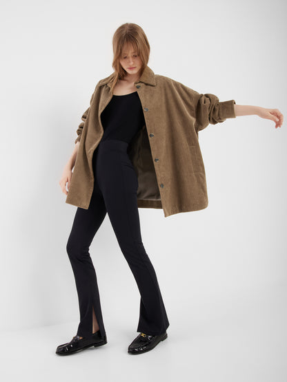 Eine Frau trägt den lockeren Mantel Ted Khaki Coderoy über einem schwarzen Oberteil, einer taillierten schwarzen Hose mit vorderen Schlitzen am Saum und schwarzen Halbschuhen, während sie vor einem schlichten weißen Hintergrund posiert.