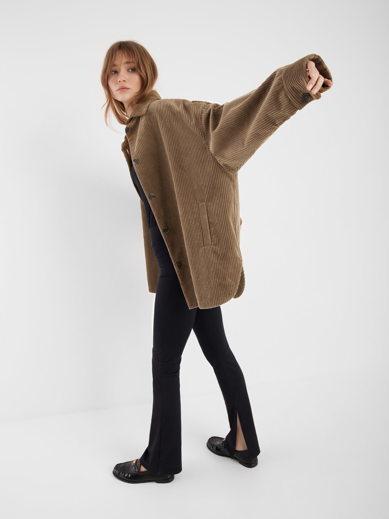 Eine Person mit hellbraunem Haar trägt den übergroßen Mantel Ted Khaki Corduroy, gepaart mit einer schwarzen Hose und schwarzen Schuhen. Sie streckt einen Arm aus und lehnt sich nach vorne vor einem schlichten weißen Hintergrund.