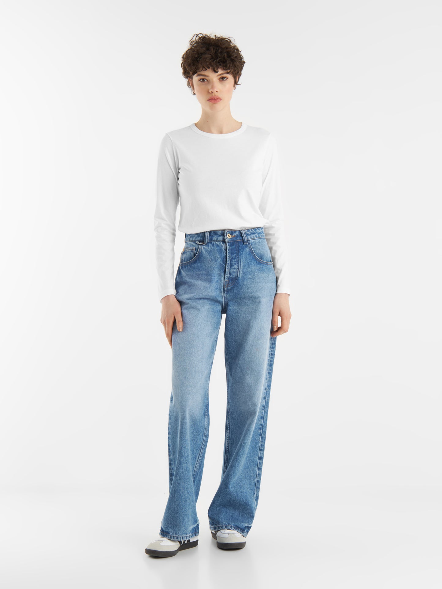 Eine Person mit kurzen lockigen Haaren trägt ein langärmeliges weißes Hemd, hochgekrempelte Bluejeans und weiße Turnschuhe und steht mit dem Gesicht nach vorne vor einem schlichten weißen Hintergrund.