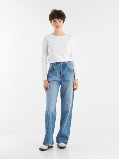 Eine Person mit kurzen lockigen Haaren trägt ein langärmeliges weißes Hemd, hochgekrempelte Bluejeans und weiße Turnschuhe und steht mit dem Gesicht nach vorne vor einem schlichten weißen Hintergrund.