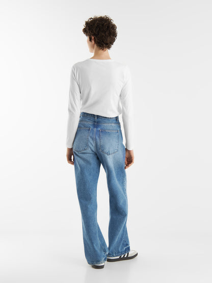 Eine Person mit kurzen lockigen Haaren, bekleidet mit einem langärmeligen weißen Oberteil, blauen Jeans und schwarz-weißen Turnschuhen, steht mit dem Gesicht zur Seite vor einem schlichten weißen Hintergrund.