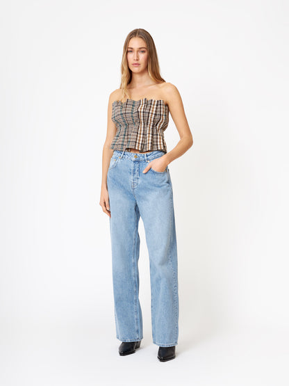 Eine Frau steht vor einem unifarbenen weißen Hintergrund und trägt ein Plissee-Top-Karo und hellblaue Jeans mit hoher Taille und weitem Bein sowie schwarze Schuhe. Sie hat langes glattes Haar und eine Hand in der Tasche.