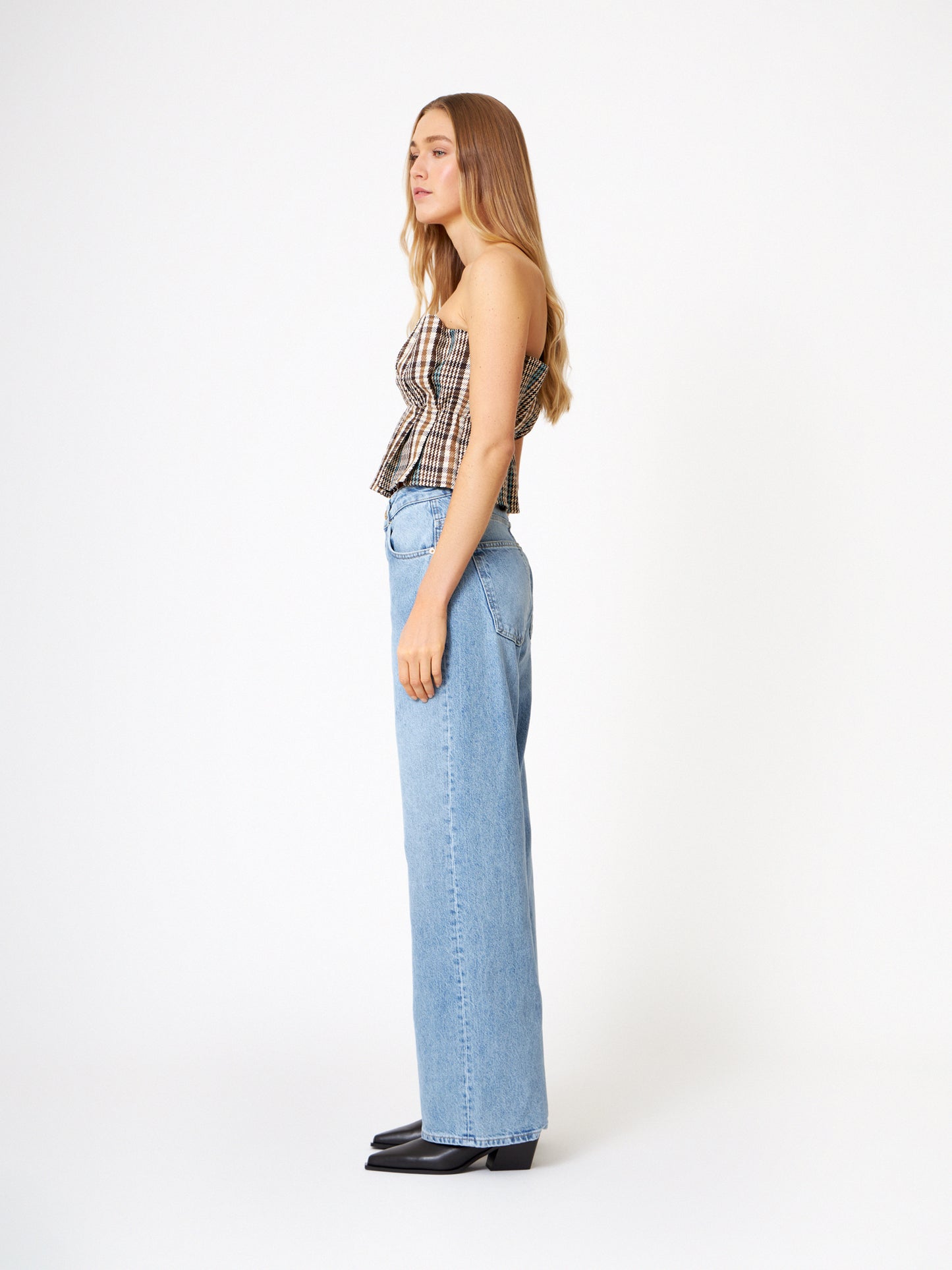 Eine Frau mit langen Haaren steht im Profil vor einem weißen Hintergrund. Sie trägt das Pleat Top Check, eine hochtaillierte Bluejeans mit weitem Bein und schwarze Schuhe.
