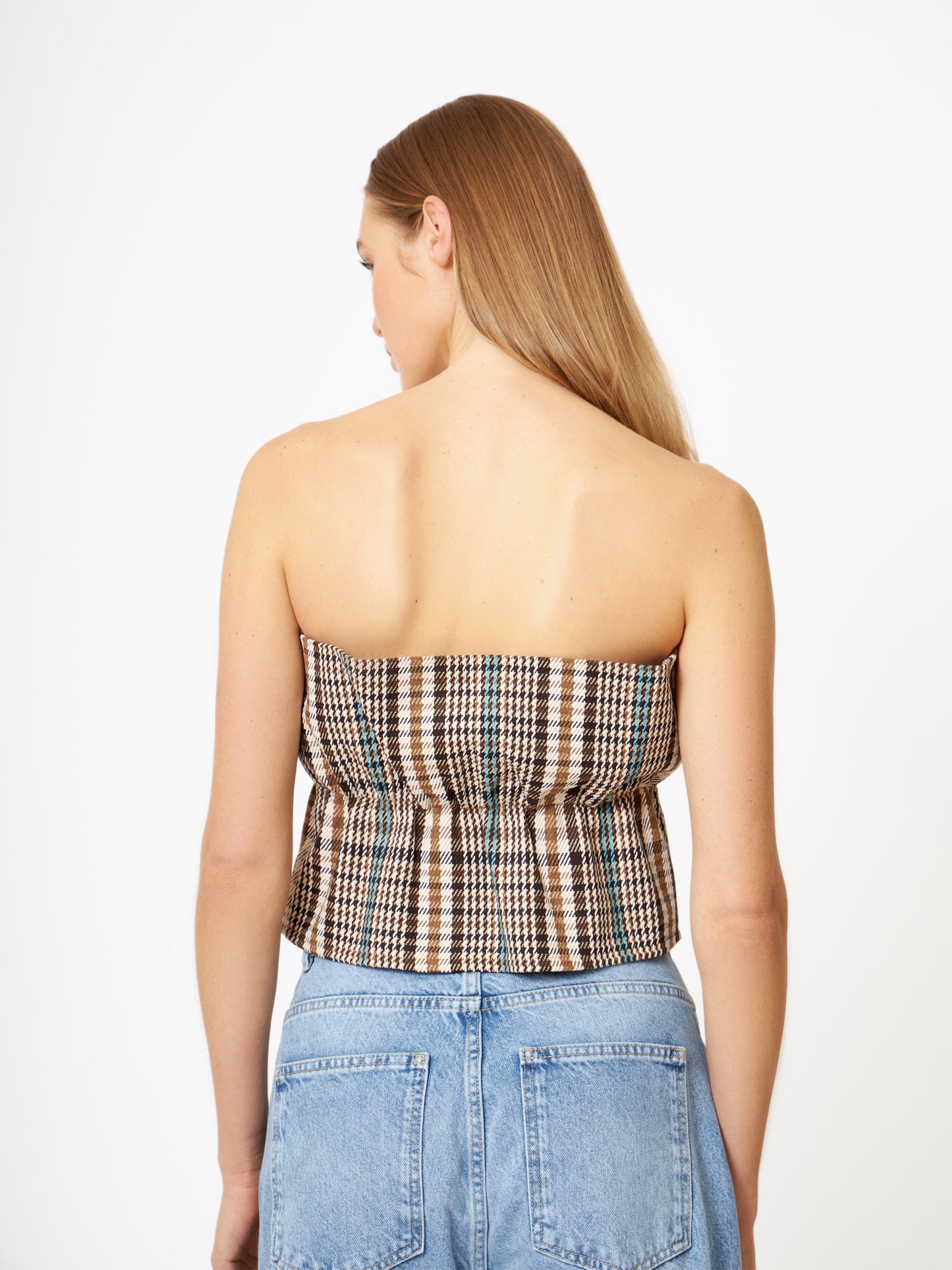 Eine Frau mit langem, glattem blondem Haar ist von hinten zu sehen. Sie trägt ein Pleat Top Check in braun-weißem Karo und hellblaue High-Waist-Jeans. Der Hintergrund ist einfarbig weiß.