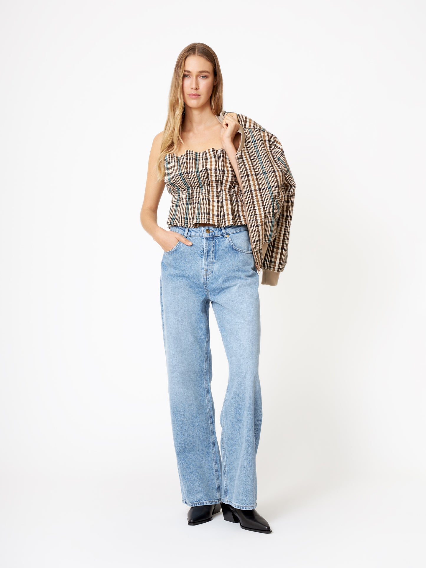 Eine Frau steht vor einem weißen Hintergrund in hellblauen Jeans mit weitem Bein, schwarzen spitzen Schuhen und dem Pleat Top Check schulterfrei Crop Top Damen und trägt eine passende karierte Jacke über einer Schulter. Sie hat lange Haare.