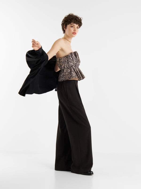 Eine Person mit kurzen lockigen Haaren trägt das trägerlose Oberteil mit Leopardenmuster von Let's Go Leo und eine weite schwarze Hose. Sie hält eine schwarze Jacke über eine Schulter und posiert selbstbewusst vor einem schlichten weißen Hintergrund.