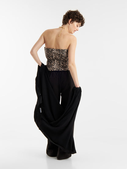 Eine Frau mit kurzen lockigen Haaren, von hinten gesehen, trägt den Pleat Top Leo und eine schwarze Hose, während sie einen langen schwarzen Mantel in den Händen hält, vor einem schlichten weißen Hintergrund.