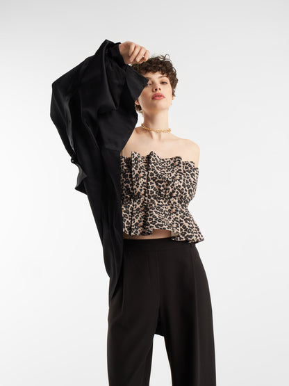 Eine Person steht vor einem unifarbenen Hintergrund, trägt eine schwarze Hose und das Pleat Top Leo, ein gerafftes Oberteil mit Leopardenmuster, schulterfrei und hält eine schwarze Jacke oder ein schwarzes Hemd in einer Hand über den Kopf.