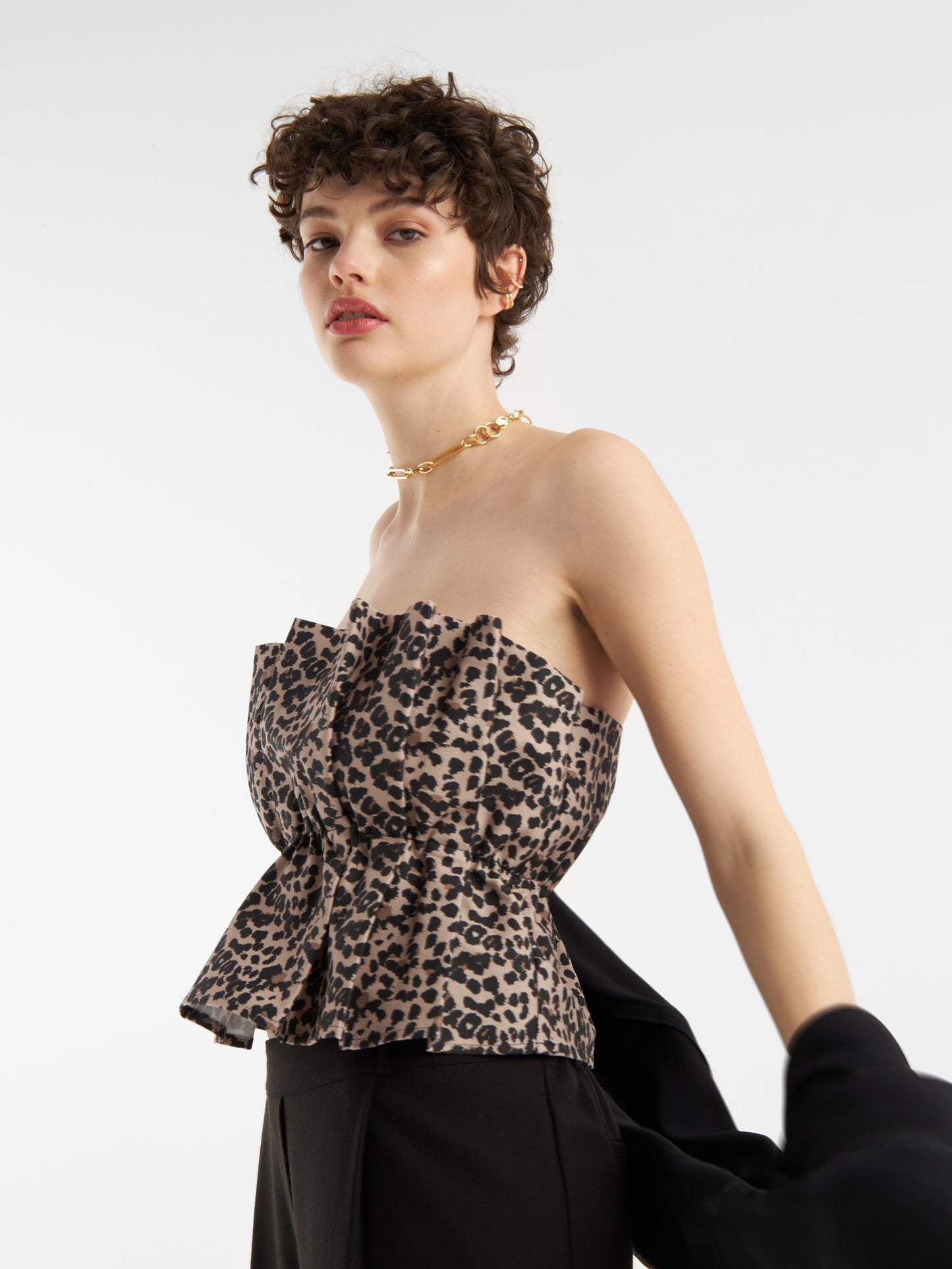Eine Frau mit kurzen lockigen Haaren trägt das Pleat Top Leo - ein schulterfreies, gerafftes Damenoberteil mit Leopardenfaltenmuster - und eine goldene Halskette und hält eine schwarze Jacke vor einem weißen Hintergrund.