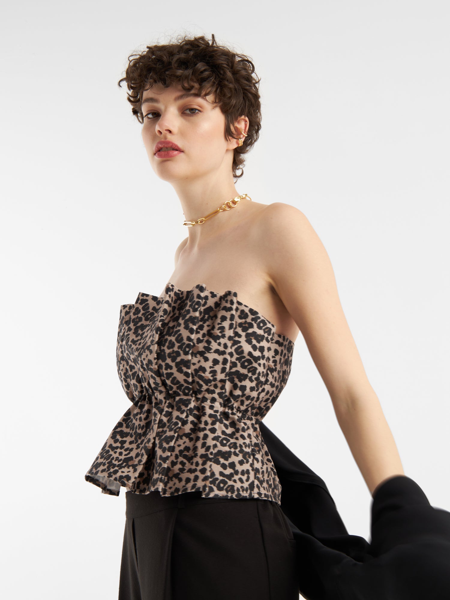Eine Frau mit kurzen lockigen Haaren trägt das Pleat Top Leo - ein schulterfreies, gerafftes Damenoberteil mit Leopardenfaltenmuster - und eine goldene Halskette und hält eine schwarze Jacke vor einem weißen Hintergrund.