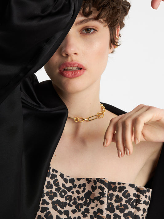 Eine Frau mit kurzen braunen Haaren posiert mit einem erhobenen Arm, der teilweise ihr Gesicht verdeckt. Sie trägt die goldene Halskette SHINE FOR CHRISTMAS! und ein trägerloses Top mit Leopardenmuster.