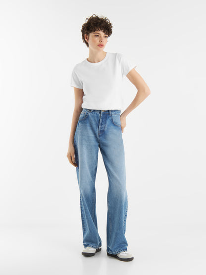 Eine Person mit kurzen, lockigen Haaren trägt ein weißes T-Shirt mit Rundhalsausschnitt, hochgekrempelte Bluejeans und weiße Turnschuhe und steht vor einem schlichten weißen Hintergrund.