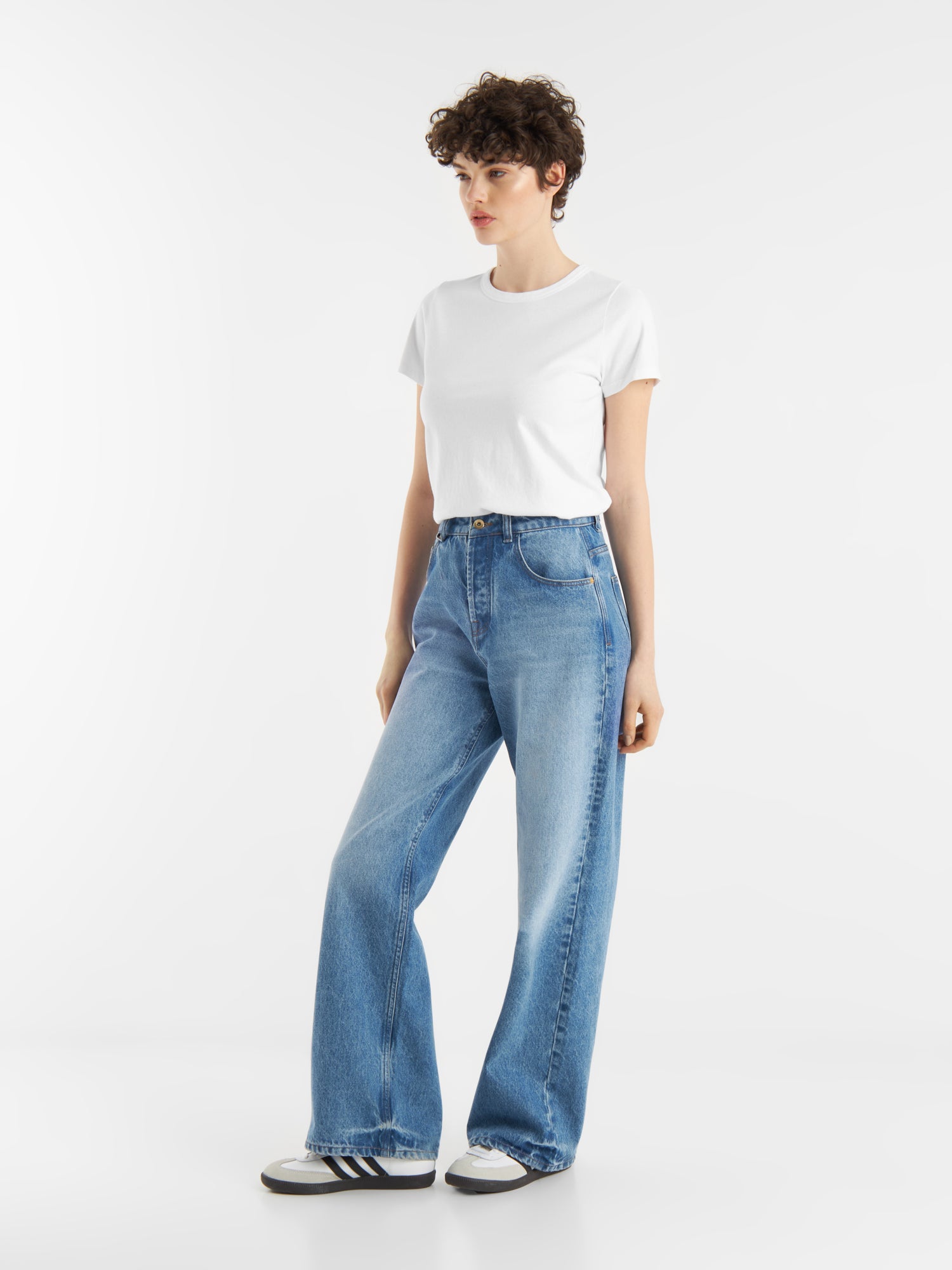 Eine Person mit kurzen lockigen Haaren trägt ein weißes T-Shirt mit Rundhalsausschnitt, hellblaue Jeans mit weitem Bein und schwarz-weiße Turnschuhe vor einem schlichten weißen Hintergrund.