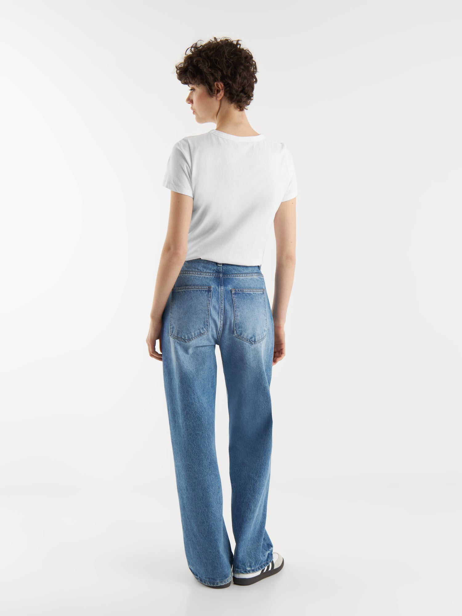 Eine Person mit kurzen lockigen Haaren trägt das T-Shirt Roundneck White, weite Bluejeans und schwarz-weiße Turnschuhe und steht mit dem Rücken zu einem schlichten weißen Hintergrund.