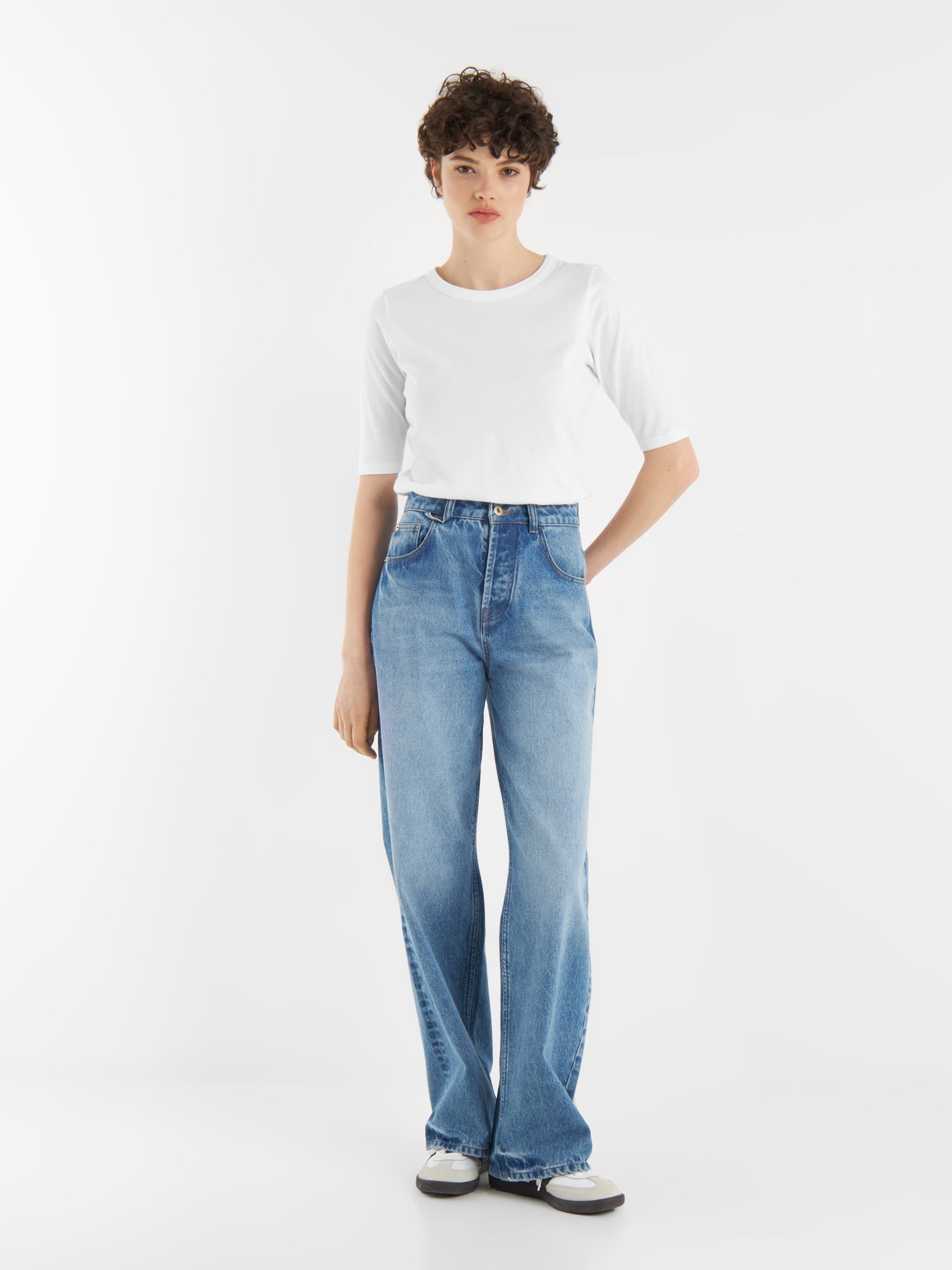 Eine Person mit kurzen lockigen Haaren trägt ein weißes T-Shirt, Jeans Pants Denim und schwarz-weiße Turnschuhe und steht vor einem schlichten weißen Hintergrund.