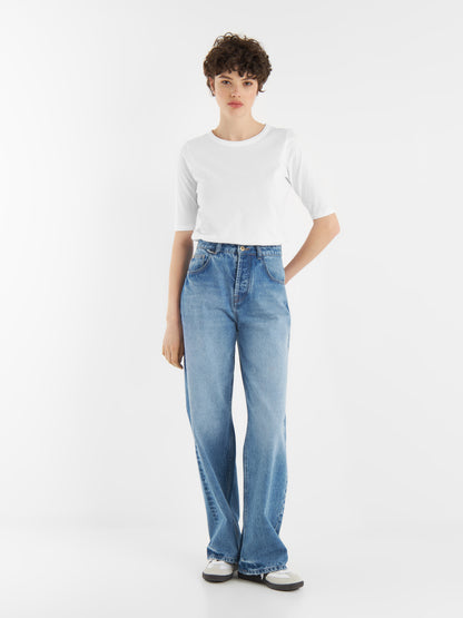 Eine Person mit kurzen lockigen Haaren trägt ein weißes T-Shirt, Jeans Pants Denim und schwarz-weiße Turnschuhe und steht vor einem schlichten weißen Hintergrund.