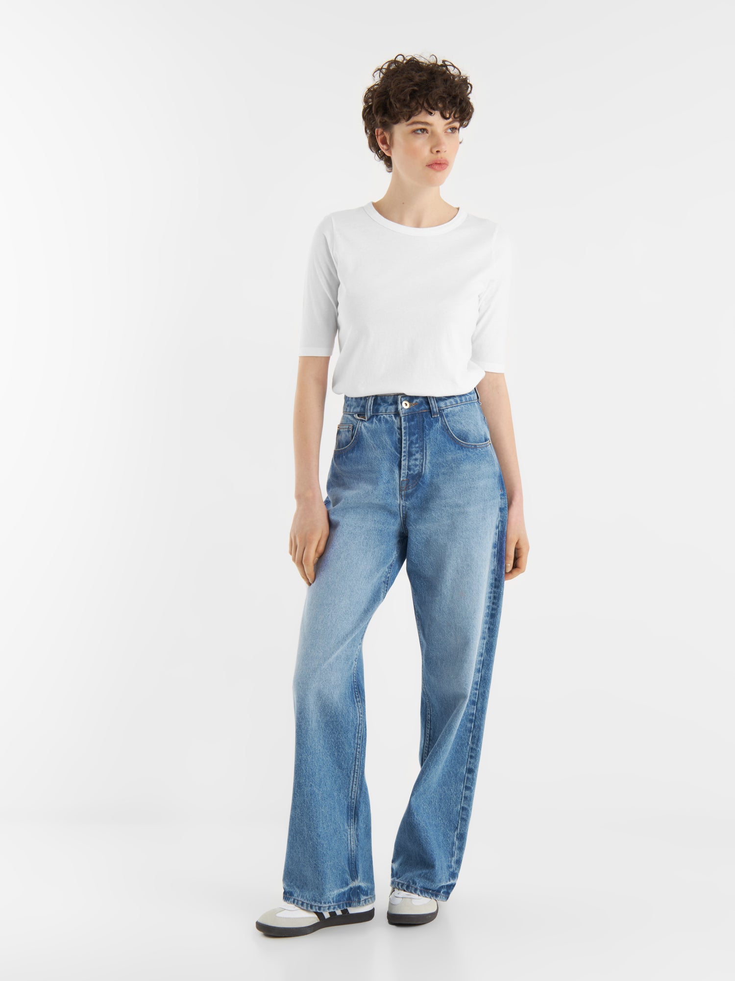 Eine Person mit kurzen lockigen Haaren trägt ein weißes T-Shirt mit halben Ärmeln, eine hochgeschnittene blaue Jeans mit weitem Bein und schwarz-weiße Schuhe und steht vor einem schlichten weißen Hintergrund.