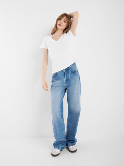 Eine Frau steht vor einem weißen Hintergrund und trägt ein schlichtes weißes T-Shirt, eine hellblaue Jeanshose mit weitem Bein und weiße Turnschuhe, wobei sie einen Arm hebt und ihre Hand auf dem Kopf ruht.