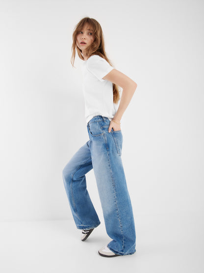 Eine Frau mit langen Haaren trägt ein weißes T-Shirt und eine hellblaue Jeanshose Denim mit schwarz-weißen Turnschuhen. Sie steht vor einem schlichten weißen Hintergrund und blickt über ihre Schulter.