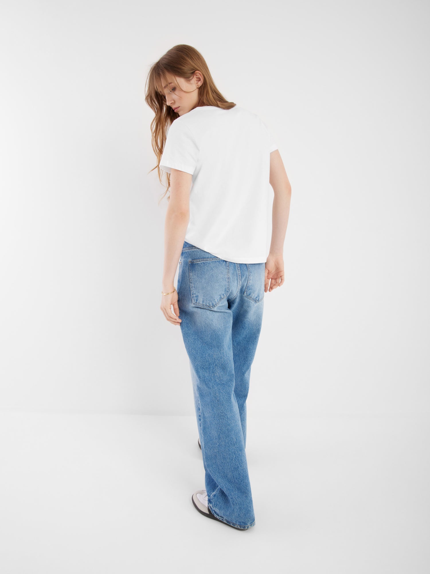 Eine Person mit langen Haaren, einem weißen T-Shirt mit V-Ausschnitt, blauen Jeans und weißen Turnschuhen steht in einem minimalistischen weißen Raum und blickt über die Schulter zurück.