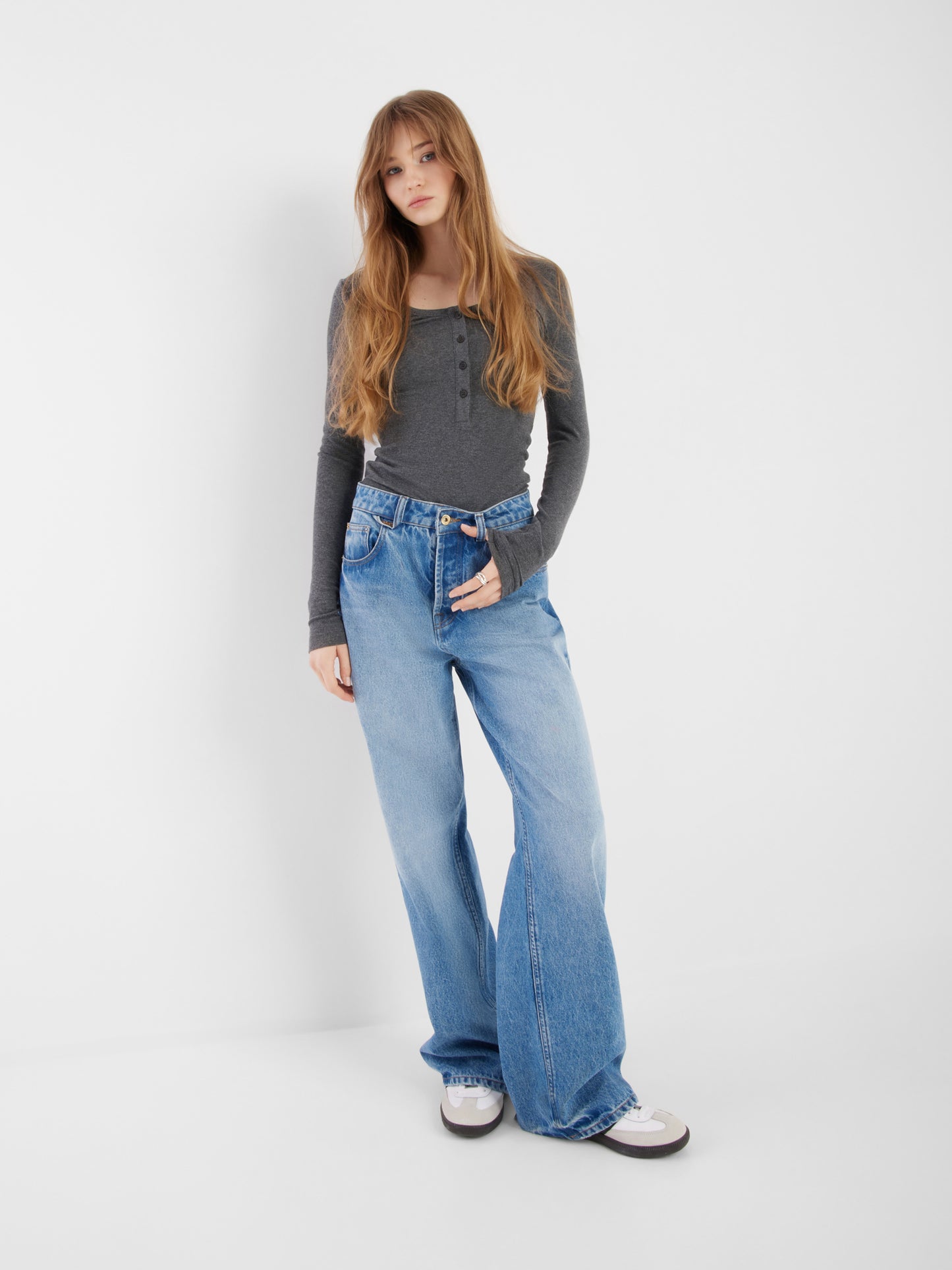 Eine junge Frau mit langem hellbraunem Haar steht vor einem schlichten weißen Hintergrund. Sie trägt ein tailliertes Oberteil aus Kaschmir mit langen Ärmeln und Knöpfen in Anthrazit, hochgekrempelte, weite Bluejeans und weiße Turnschuhe. Sie hat eine Hand in der Tasche und sieht entspannt aus.