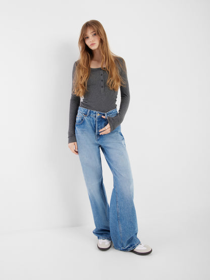 Eine junge Frau mit langem hellbraunem Haar steht vor einem schlichten weißen Hintergrund. Sie trägt ein tailliertes Oberteil aus Kaschmir mit langen Ärmeln und Knöpfen in Anthrazit, hochgekrempelte, weite Bluejeans und weiße Turnschuhe. Sie hat eine Hand in der Tasche und sieht entspannt aus.