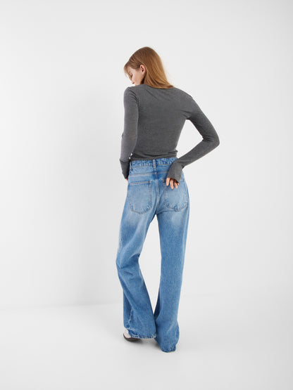 Eine Frau mit glattem hellbraunem Haar trägt ein tailliertes Oberteil aus Kaschmir mit langen Ärmeln und Knöpfen in Anthrazit und eine hochgeschnittene blaue Jeans mit weitem Bein. Sie steht mit einer Hand in der Gesäßtasche vor einem schlichten weißen Hintergrund.