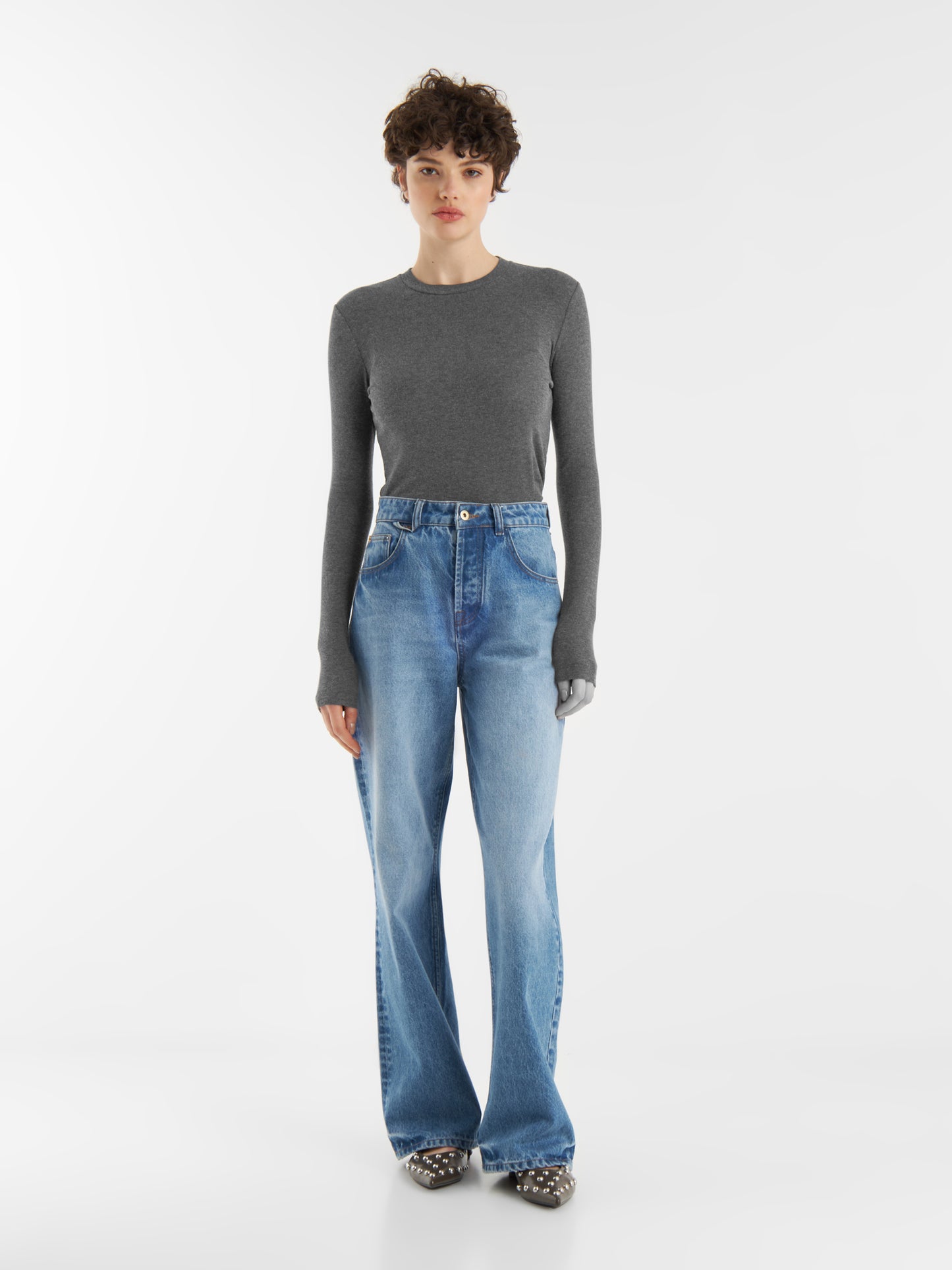 Eine Person mit kurzen lockigen Haaren trägt einen taillierten Kaschmir-Langarm-Anthrazit, hochgekrempelte blaue Jeans mit weitem Bein und gepunktete Schuhe vor einem schlichten weißen Hintergrund.