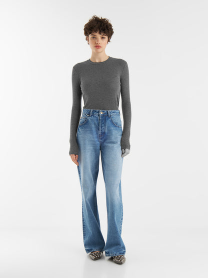 Eine Person mit kurzen lockigen Haaren trägt einen taillierten Kaschmir-Langarm-Anthrazit, hochgekrempelte blaue Jeans mit weitem Bein und gepunktete Schuhe vor einem schlichten weißen Hintergrund.