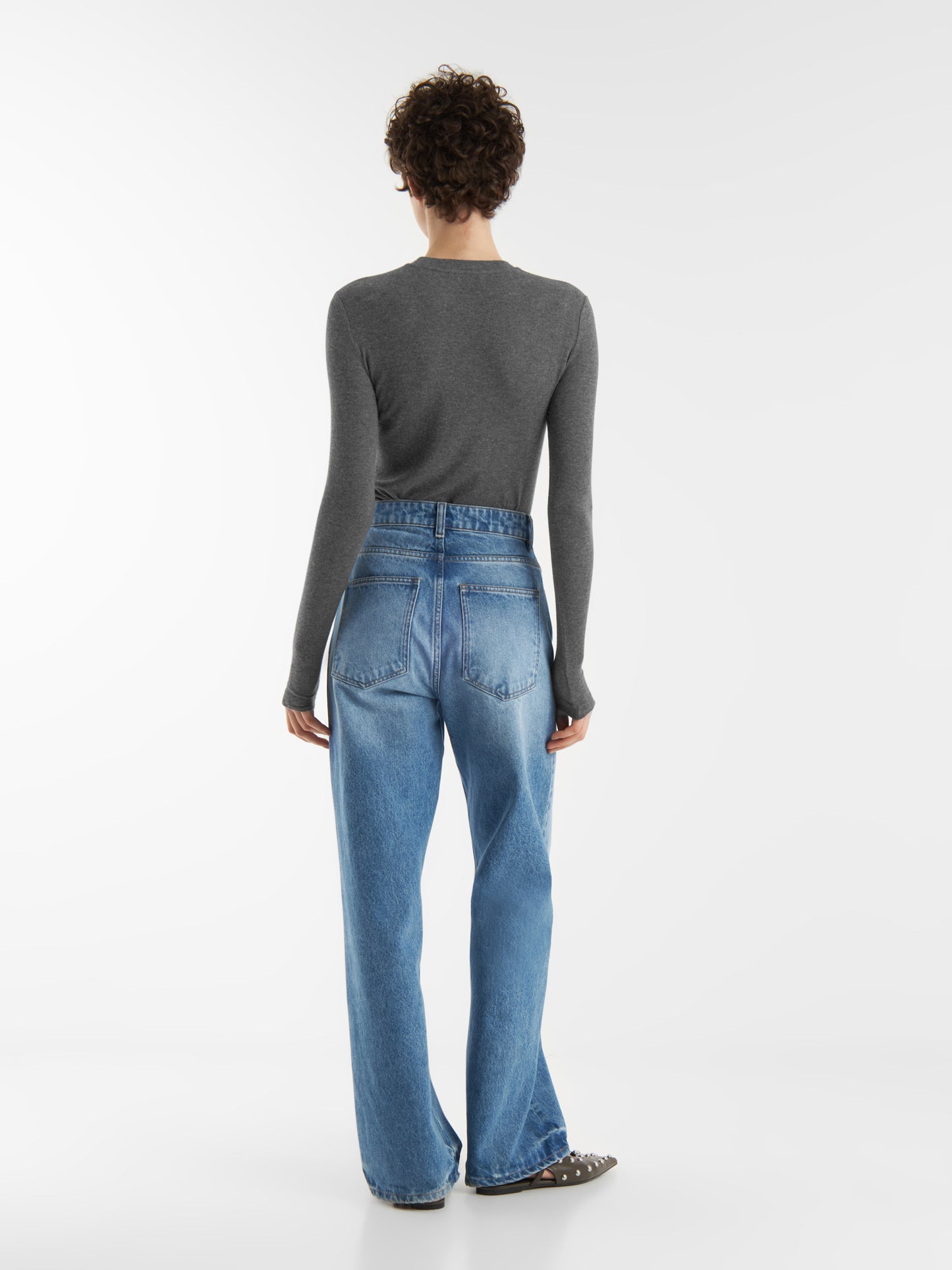 Person, die mit dem Rücken zur Kamera steht und einen grauen, taillierten Kaschmir-Langarm-Anthrazit, Bluejeans mit hoher Taille und flache Schuhe vor einem weißen Hintergrund trägt.