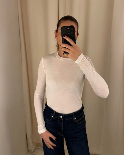 Eine Person, die das Oberteil Cashmere Long Sleeve Rib White und dunkle Jeans mit hoher Taille trägt, macht ein Spiegel-Selfie mit einem schwarzen Telefon, das Gesicht verdeckt. Im Hintergrund sind beigefarbene Vorhänge zu sehen.
