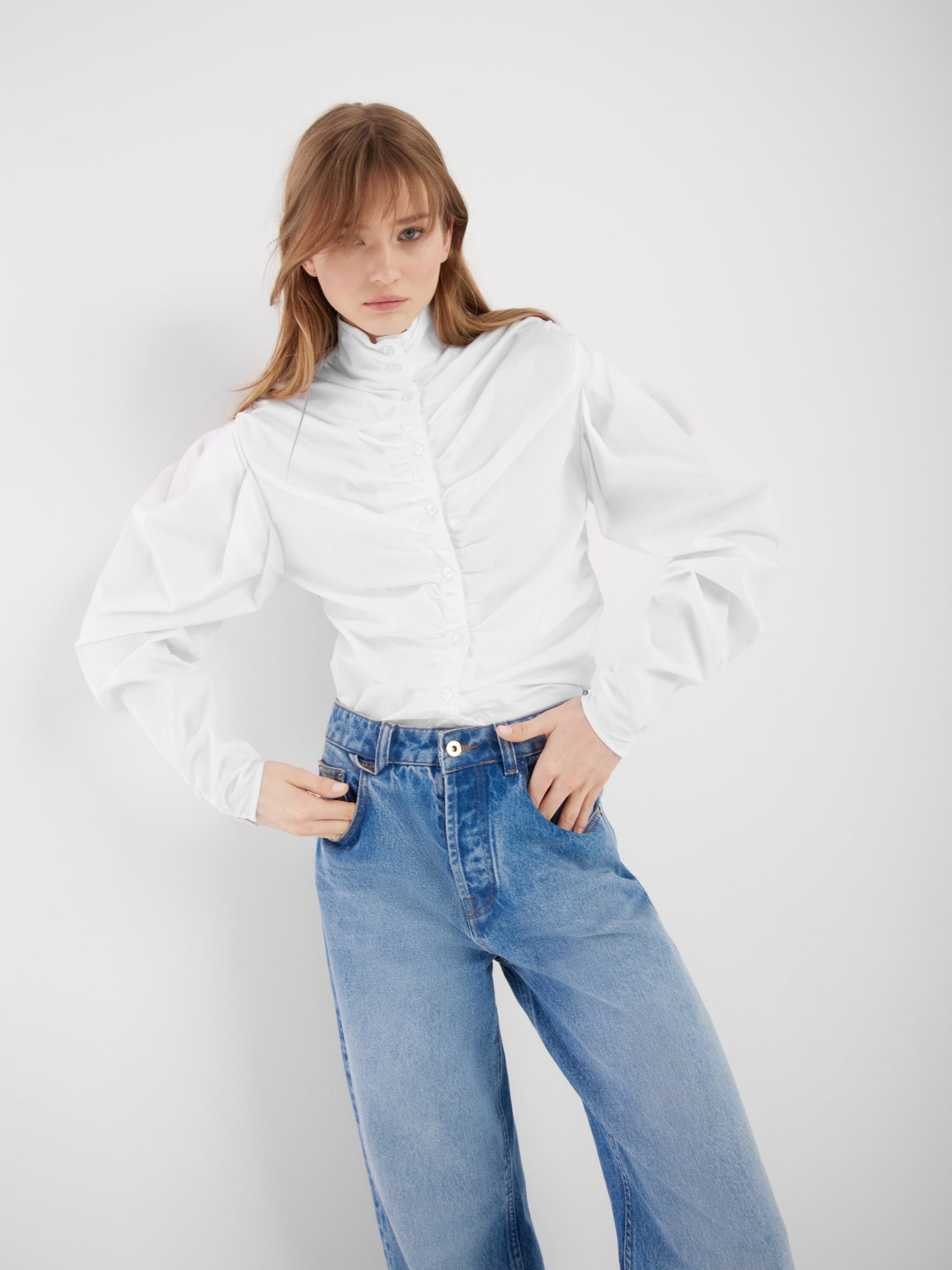 Eine Frau steht vor einem weißen Hintergrund und trägt die Bluse Virginy weiß mit Stehkragen, gerüschten Details, Puffärmeln und hellblauen, hoch sitzenden Jeans. Sie blickt in die Kamera und hat die Hände in die Hüften gestemmt.