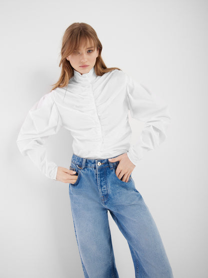 Eine Frau steht vor einem weißen Hintergrund und trägt die Bluse Virginy weiß mit Stehkragen, gerüschten Details, Puffärmeln und hellblauen, hoch sitzenden Jeans. Sie blickt in die Kamera und hat die Hände in die Hüften gestemmt.
