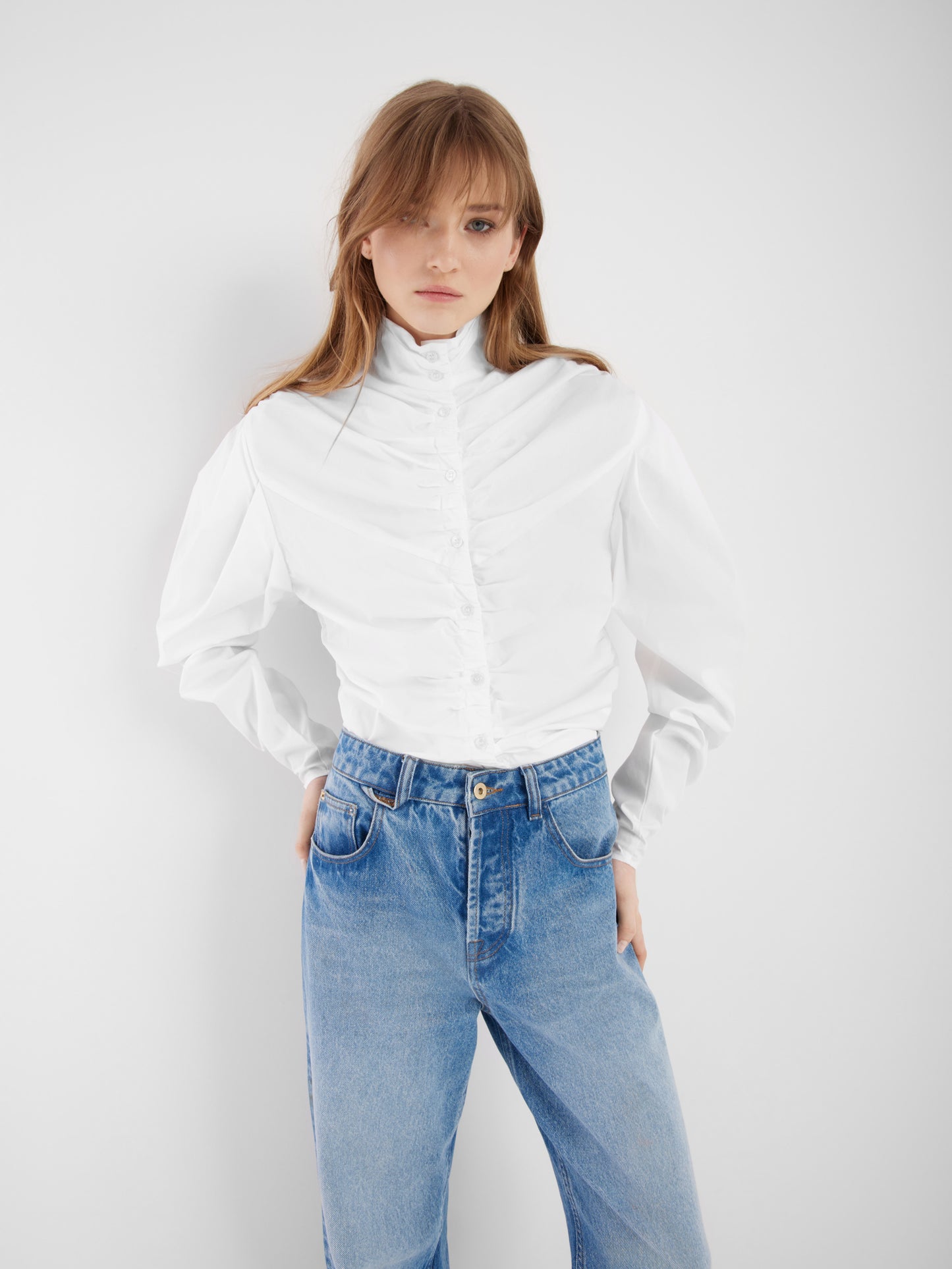 Eine Frau steht vor einem einfarbig weißen Hintergrund und trägt die Bluse Virginy weiß mit Stehkragen und Puffärmeln, gepaart mit einer blauen Jeans. Ihre Hände stecken in den Hosentaschen und ihr Haar fällt locker über ihre Schultern.