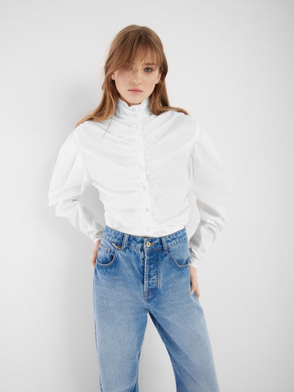 Eine Frau steht vor einem einfarbig weißen Hintergrund und trägt die Bluse Virginy weiß mit Stehkragen und Puffärmeln, gepaart mit einer blauen Jeans. Ihre Hände stecken in den Hosentaschen und ihr Haar fällt locker über ihre Schultern.