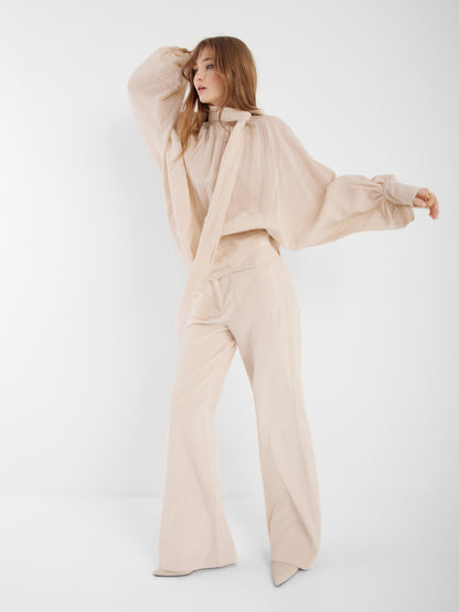 Eine Frau posiert vor einem schlichten weißen Hintergrund und trägt die "Bluse Bombay beige" mit Krawattenausschnitt und Ballonärmeln, gepaart mit einer weiten beigen Hose und spitzen Schuhen.
