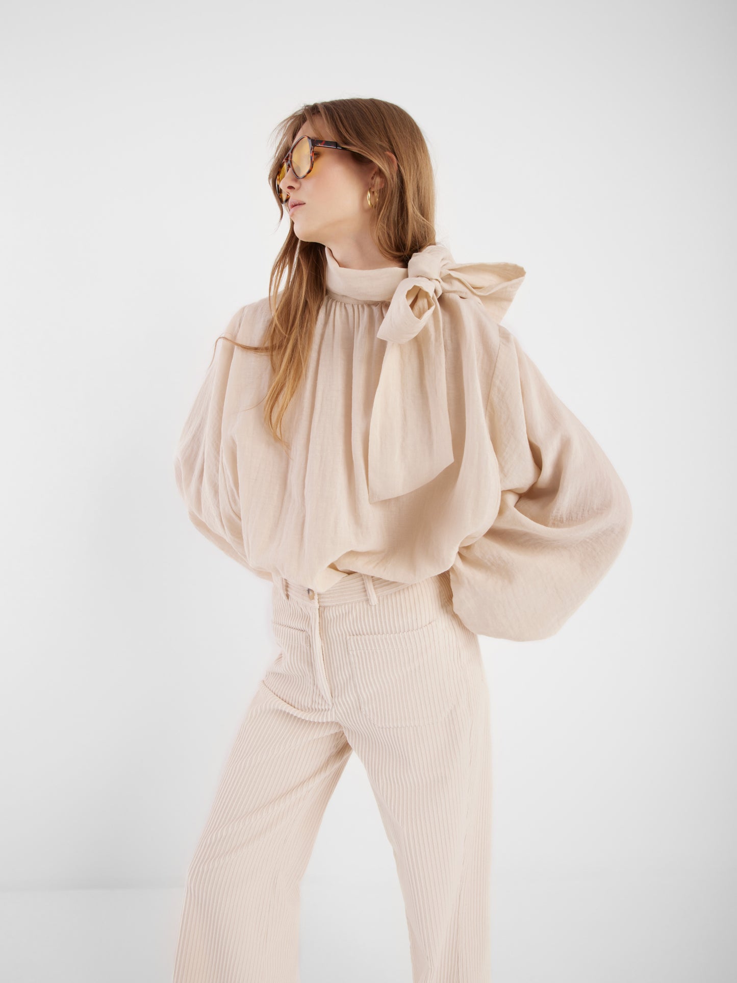 Eine Frau trägt die Bluse Bombay beige mit Schleifenkragen und passenden, hoch taillierten Hosen, steht vor weißem Hintergrund und blickt zur Seite.