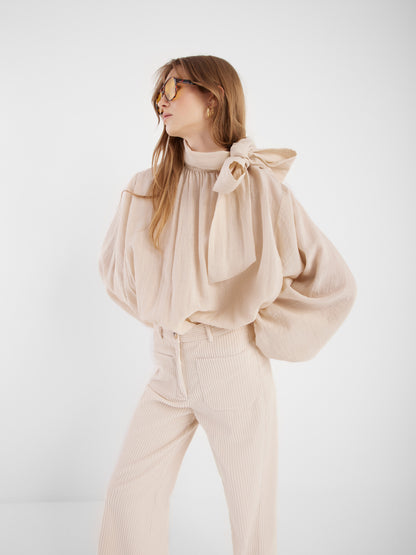 Eine Frau trägt die Bluse Bombay beige mit Schleifenkragen und passenden, hoch taillierten Hosen, steht vor weißem Hintergrund und blickt zur Seite.