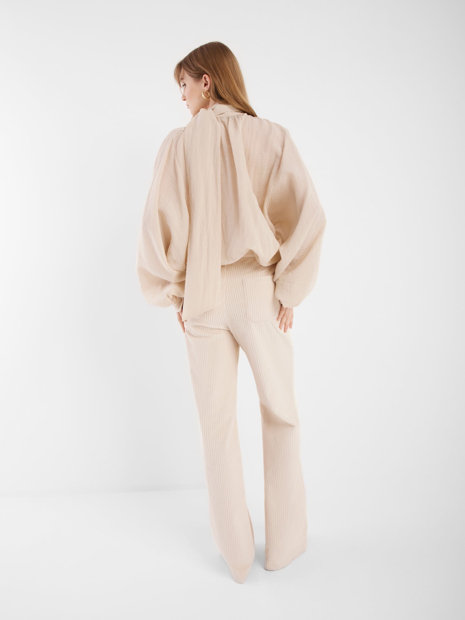Eine Frau steht mit dem Gesicht zur Wand und trägt die Bluse Bombay beige mit Ballonärmeln und passenden, weiten Hosen. Der schlichte weiße Hintergrund unterstreicht die Eleganz ihres neutral gehaltenen Outfits.