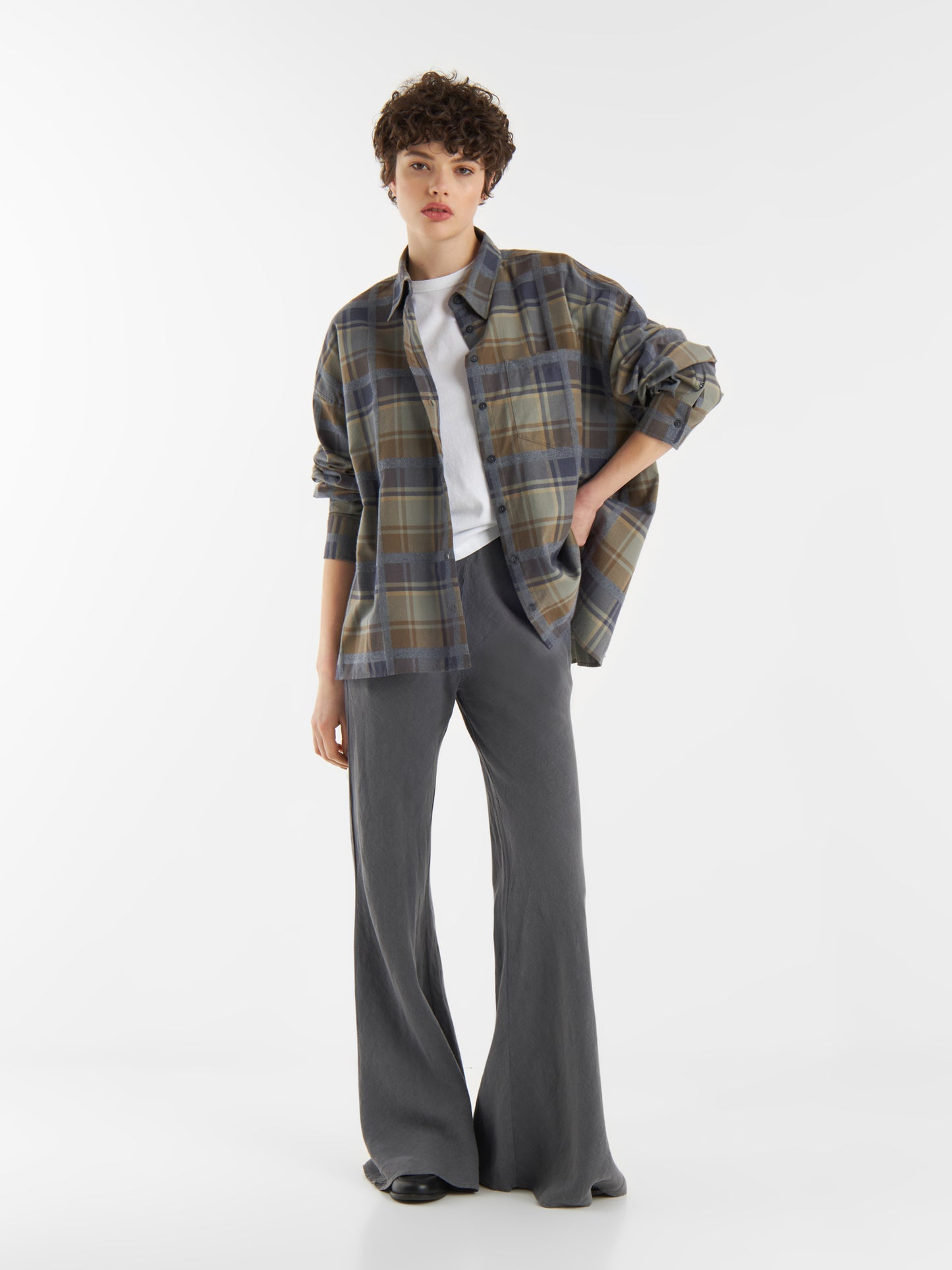 Eine Person mit kurzen lockigen Haaren trägt eine Bluse Boxy Olive über einem weißen T-Shirt und einer grauen Hose mit weitem Bein und steht mit einer Hand in der Tasche vor einem schlichten weißen Hintergrund.