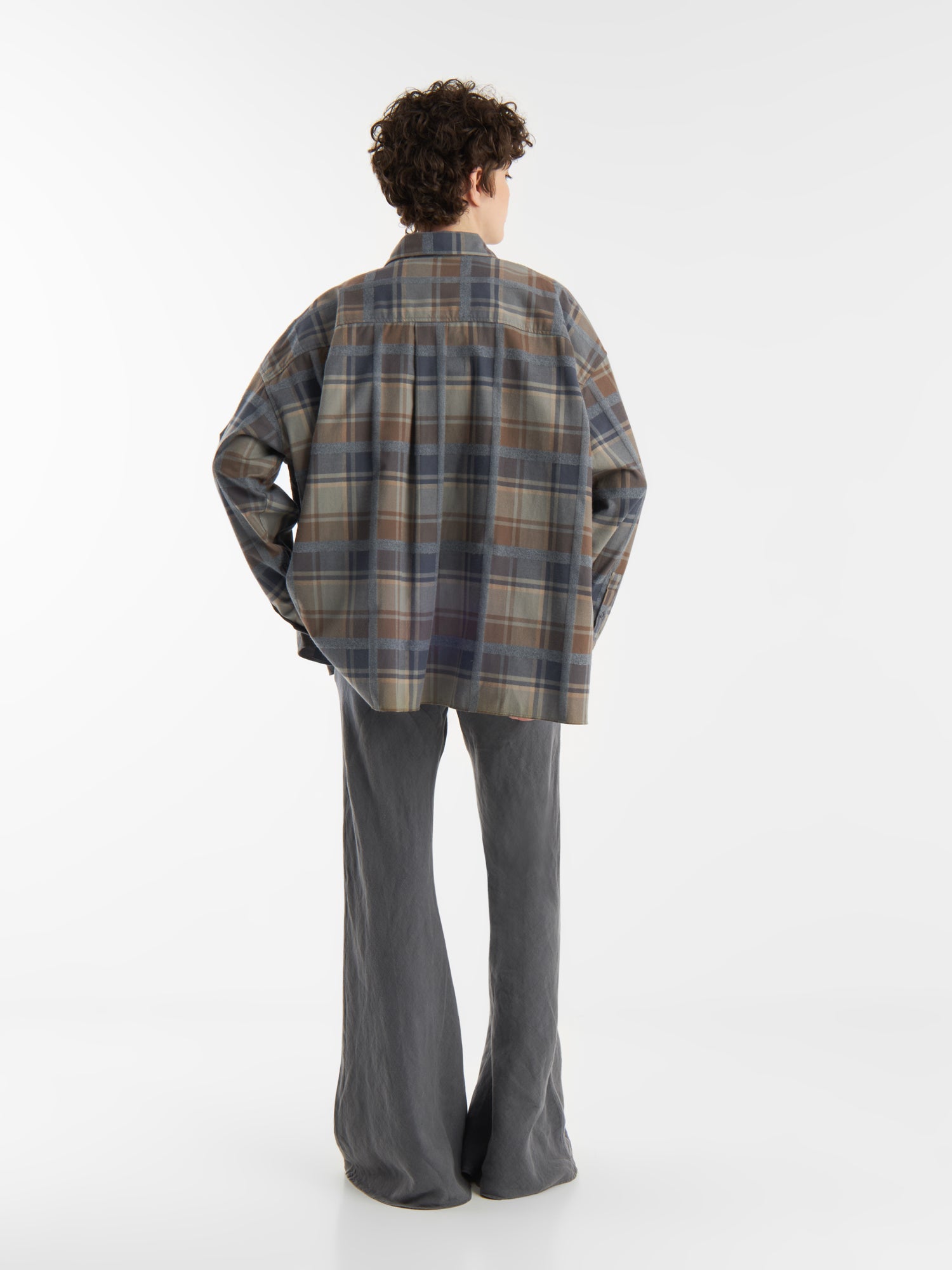 Eine Person mit kurzen, lockigen Haaren steht mit dem Gesicht zur Wand und trägt eine Bluse Boxy Olive in gedeckten Tönen und eine locker sitzende graue Hose vor einem schlichten weißen Hintergrund.