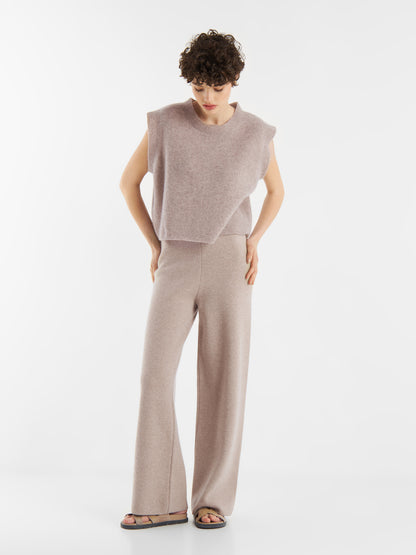 Eine Person mit kurzen lockigen Haaren trägt das Pullunder Cashmere Beige und eine dazu passende Hose mit weitem Bein. Sie steht vor einem schlichten weißen Hintergrund und schaut mit den Händen in den Hüften leicht nach unten.