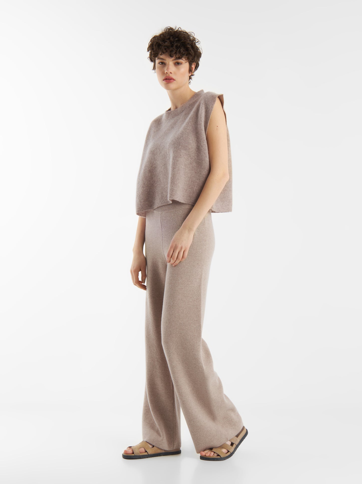 Eine Person mit kurzen lockigen Haaren trägt das Pullunder Cashmere Beige und eine dazu passende Hose mit weitem Bein und Sandalen, die vor einem schlichten weißen Hintergrund stehen.