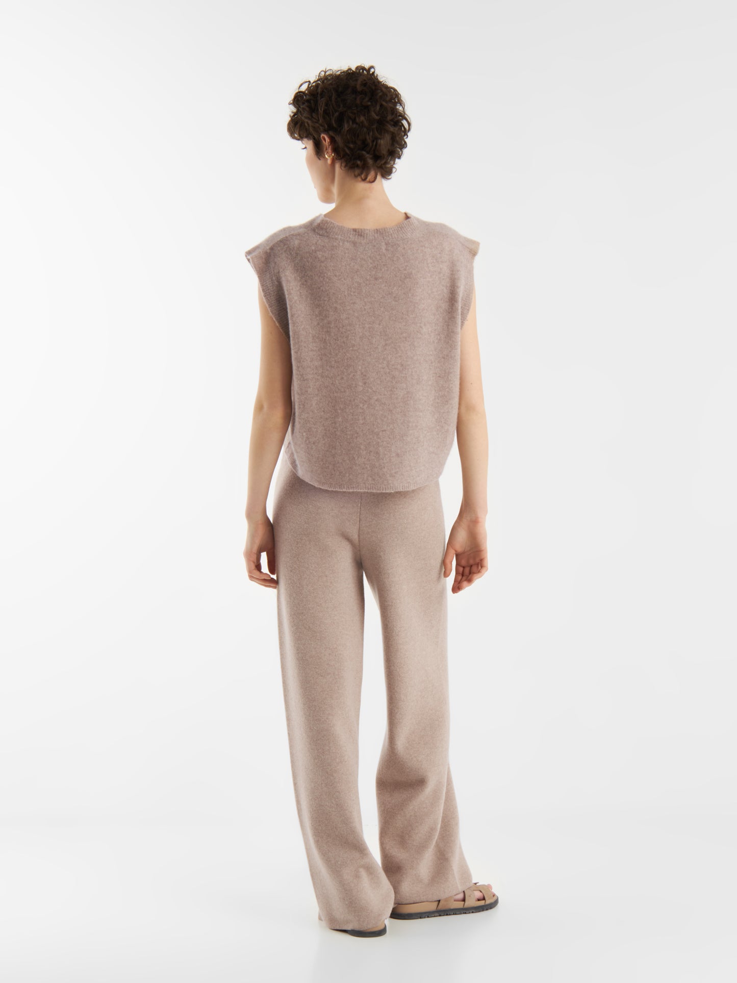 Eine Person mit kurzen lockigen Haaren blickt nach vorne und trägt das Pullunder Cashmere Beige und eine passende Hose mit weitem Bein. Das weiche, bequeme Outfit ist ideal für Pullunder Damen Styles und hebt sich von einem schlichten weißen Hintergrund ab.