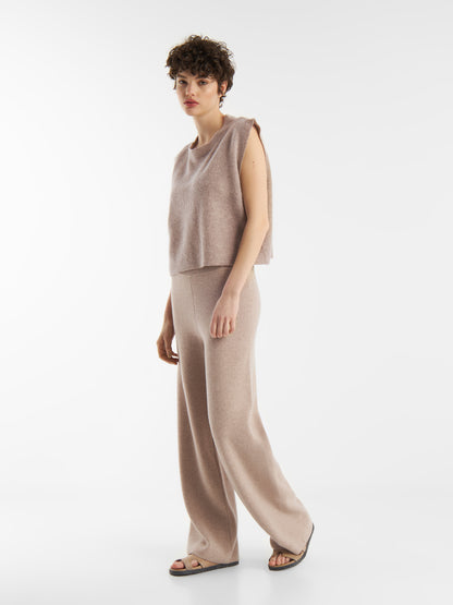 Eine Person mit kurzen lockigen Haaren trägt das Pullunder Cashmere Beige, ein ärmelloses, locker sitzendes beiges Oberteil mit passender Hose mit weitem Bein und schwarzen Sandalen, vor einem schlichten weißen Hintergrund.
