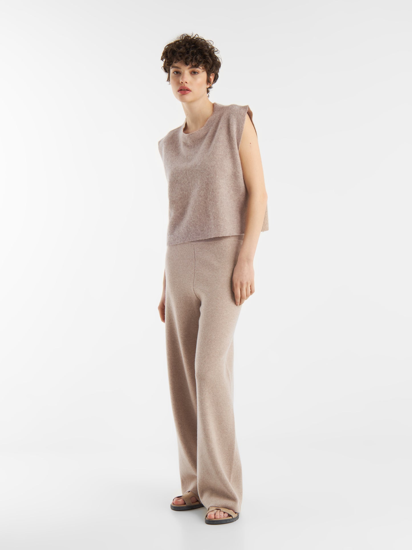 Eine Person mit kurzen lockigen Haaren trägt den Pullunder Cashmere Beige - einen ärmellosen Pullover - zusammen mit einer passenden Hose mit weitem Bein und Sandalen vor einem schlichten weißen Hintergrund.