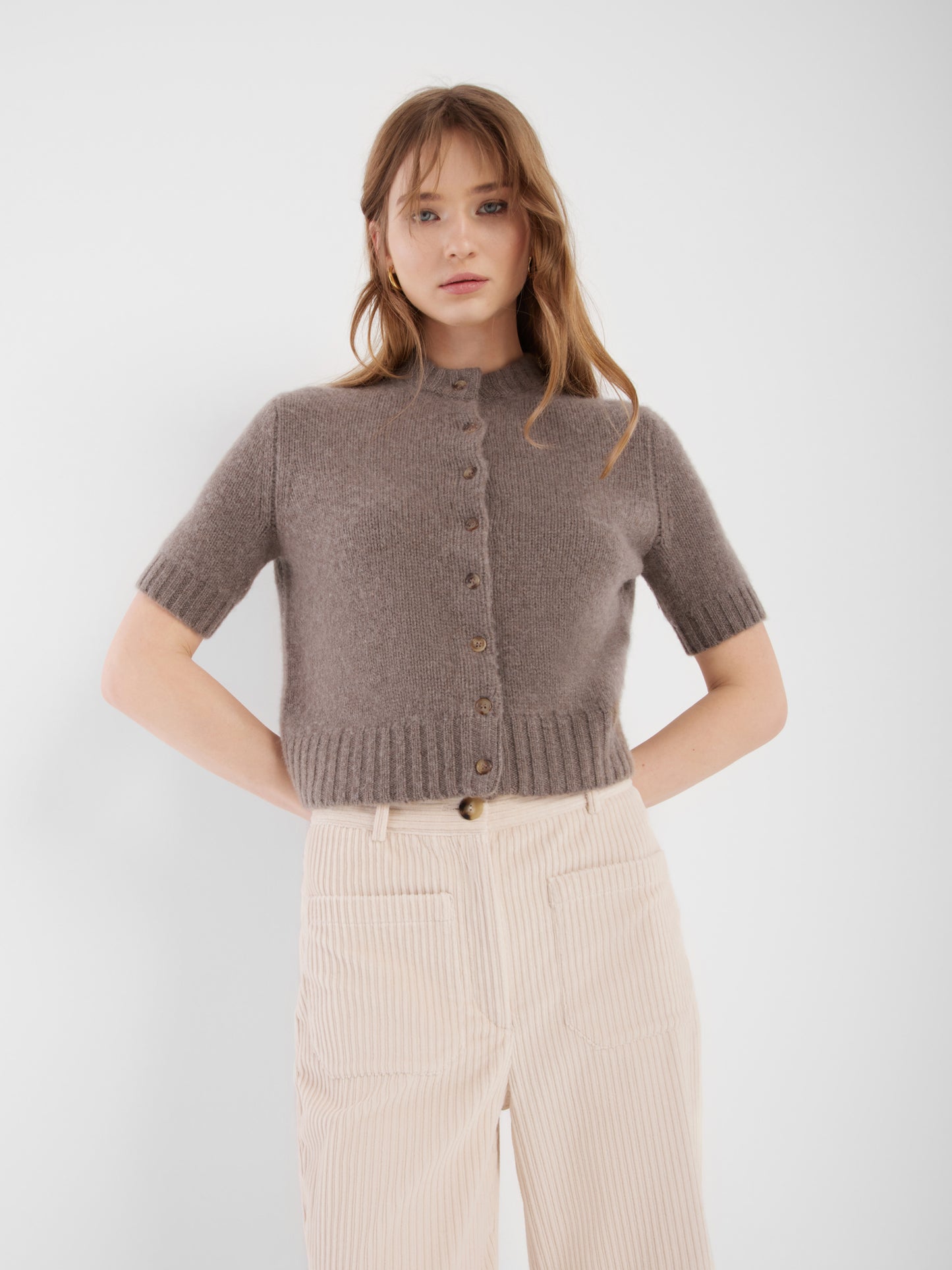 Eine Frau mit hellbraunem Haar trägt einen kurzärmeligen Azteken-Pullover in Taupebraun und eine hochtaillierte, hellbeige Hose. Sie steht vor einem schlichten weißen Hintergrund und hat die Hände in die Hüften gestemmt.